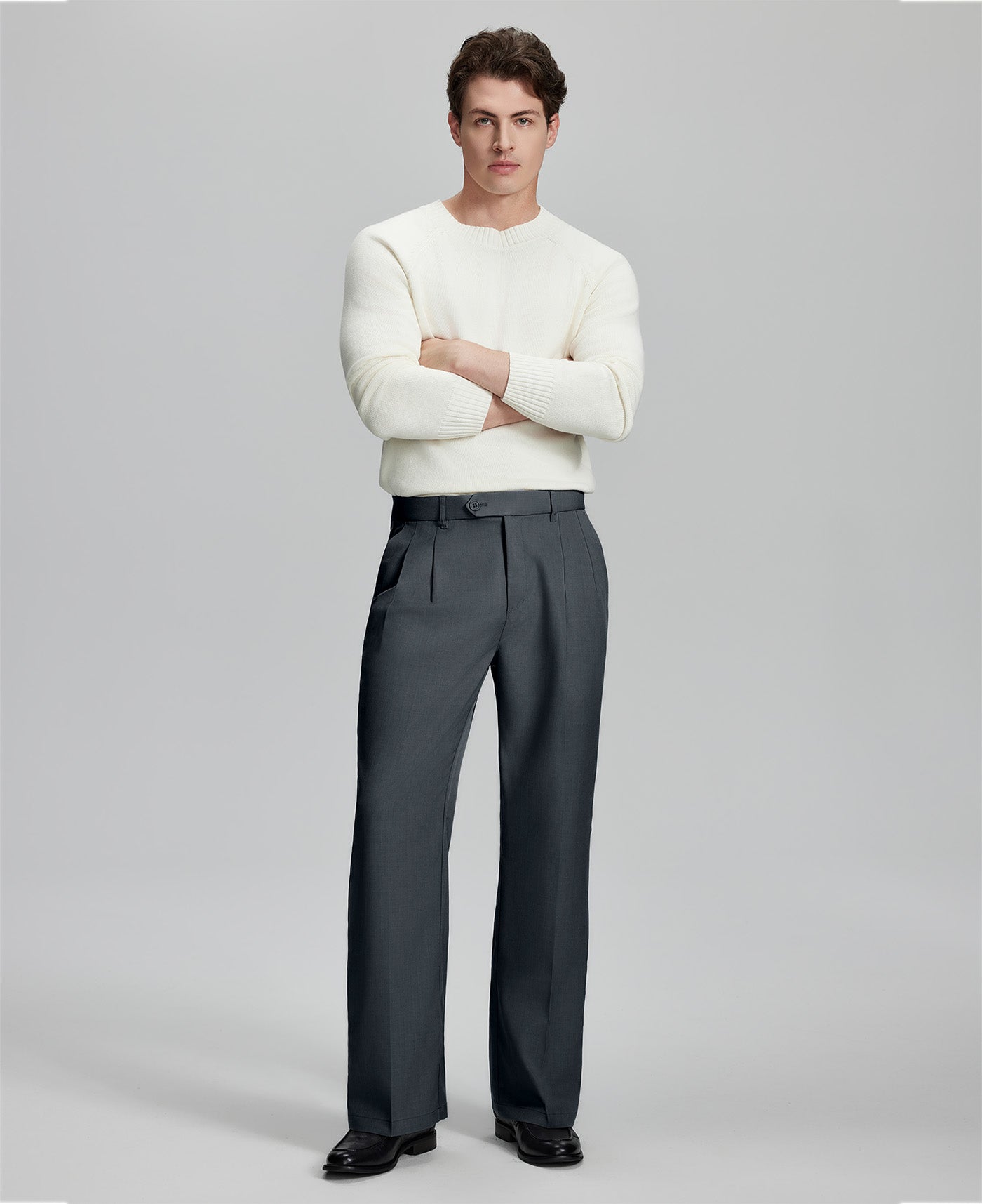 Model in white sweater, dark grey trousers & black shoes, arms crossed, presenting a smart-casual style with Baggy Pleated Dress Pants for Men.