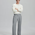 Model in white sweater, light grey trousers & black shoes, arms crossed, presenting a smart-casual style with Baggy Pleated Dress Pants for Men.