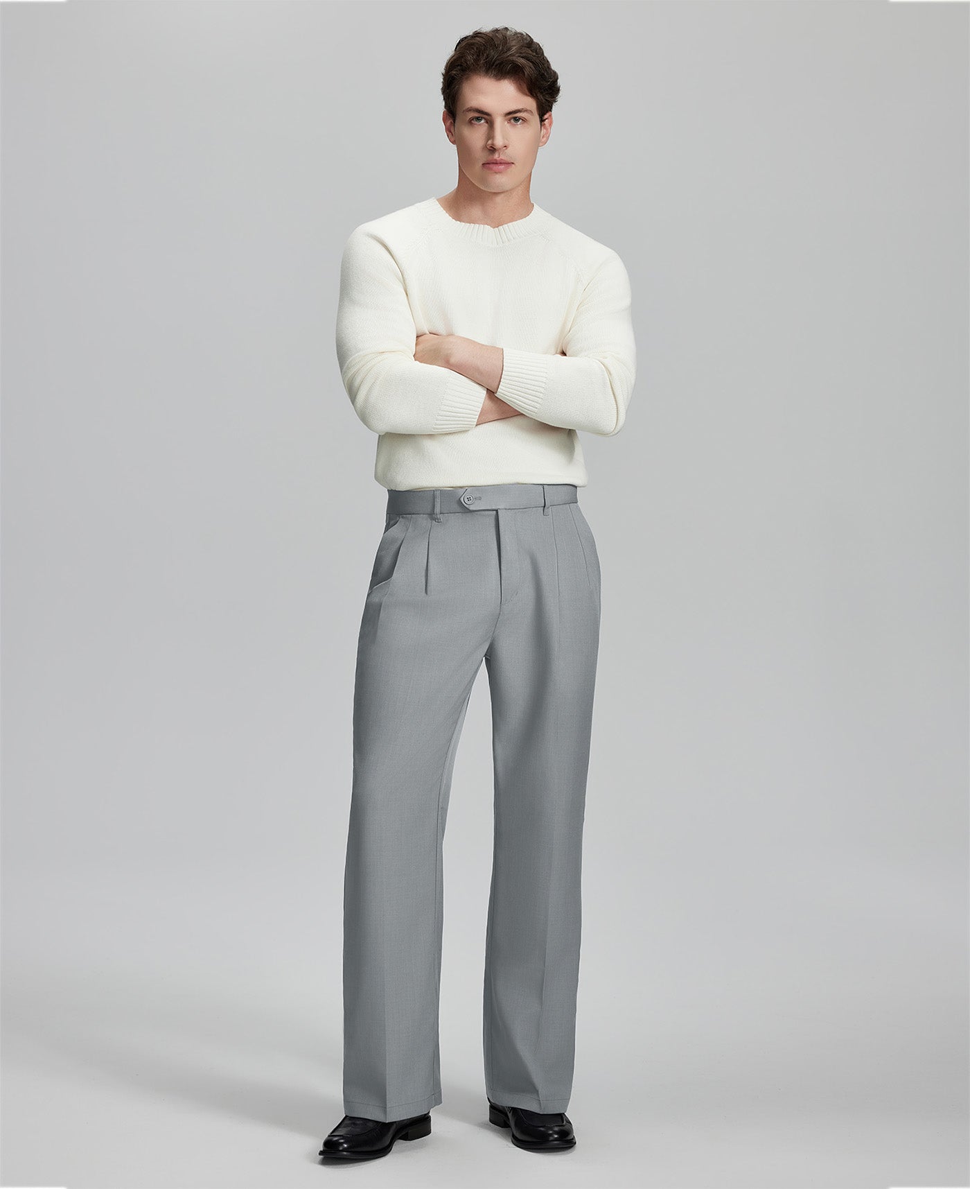 Model in white sweater, light grey trousers & black shoes, arms crossed, presenting a smart-casual style with Baggy Pleated Dress Pants for Men.