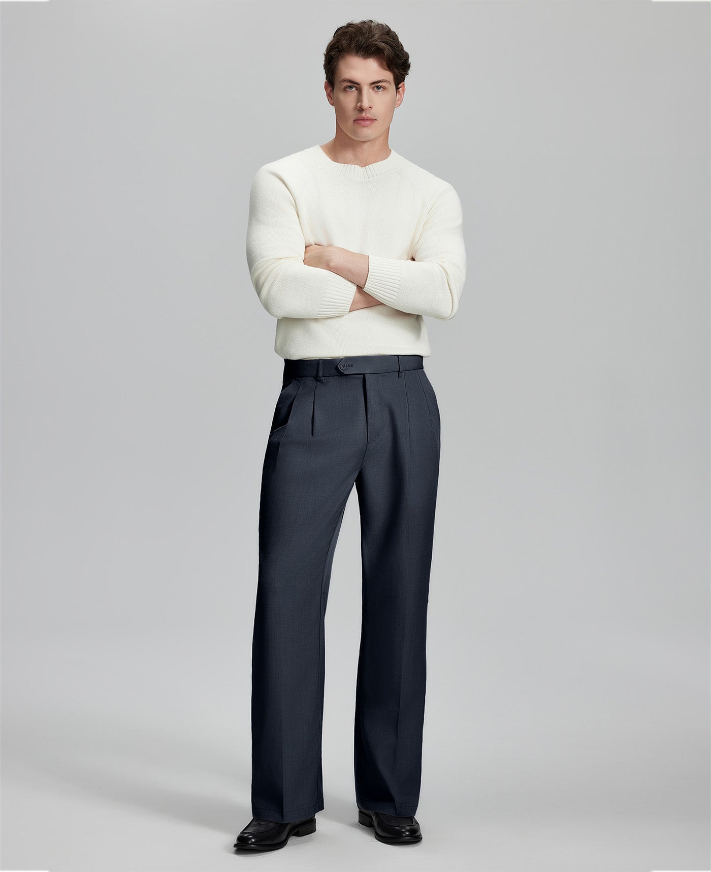 Model in white sweater, navy blue trousers & black shoes, arms crossed, presenting a smart-casual style with Baggy Pleated Dress Pants for Men.