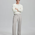 Model in white sweater, stone grey trousers & black shoes, arms crossed, presenting a smart-casual style with Baggy Pleated Dress Pants for Men.