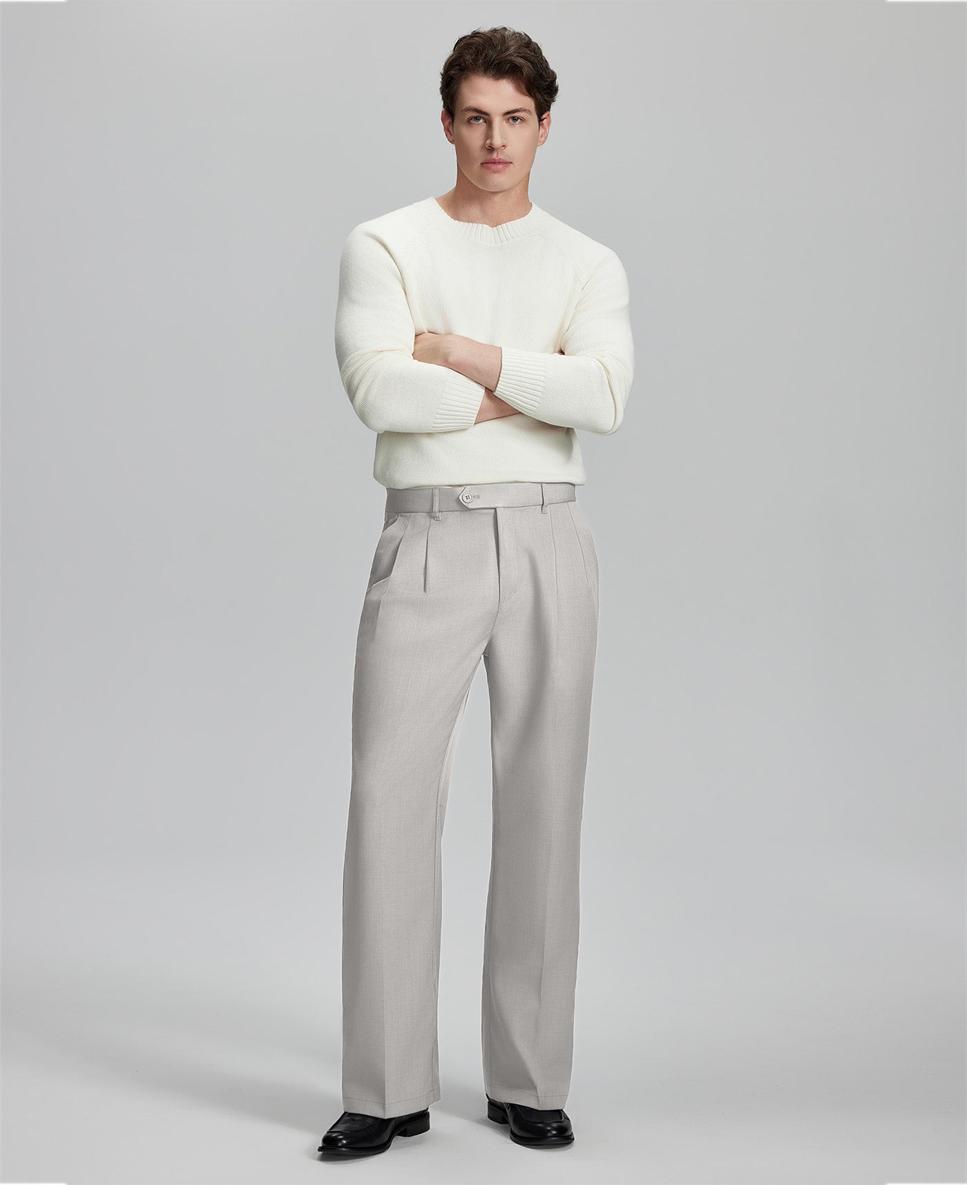 Model in white sweater, stone grey trousers & black shoes, arms crossed, presenting a smart-casual style with Baggy Pleated Dress Pants for Men.