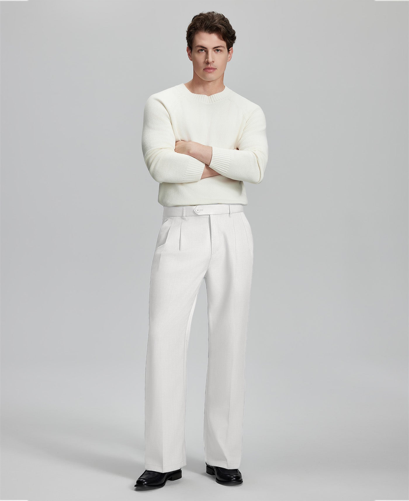 Model in white sweater, white trousers & black shoes, arms crossed, presenting a smart-casual style with Baggy Pleated Dress Pants for Men.