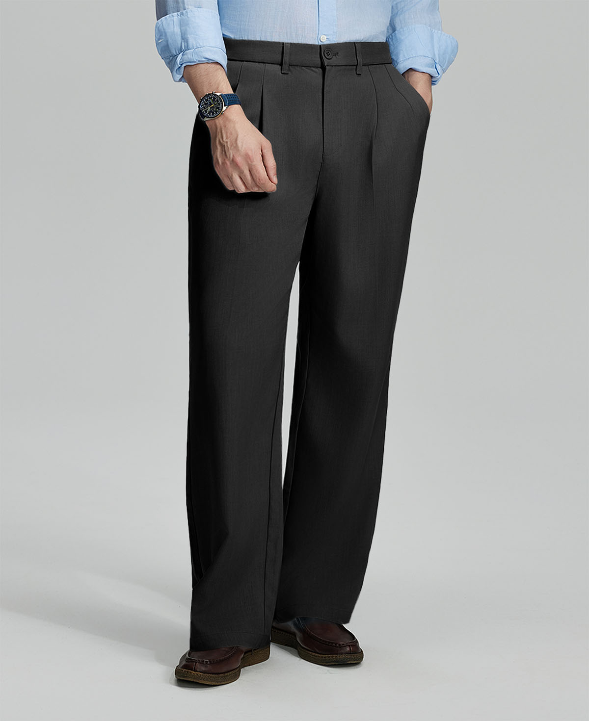 Man in light blue shirt, black pleated pants, and brown shoes, hand in pocket showcasing relaxed fit and drape.