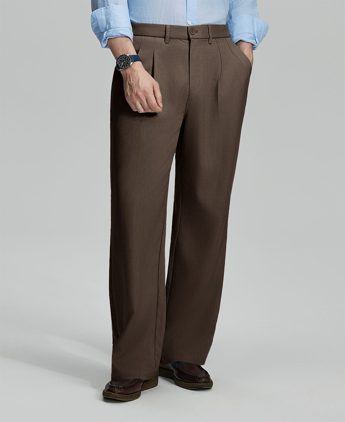 Man in light blue shirt, coffee pleated pants, and brown shoes, hand in pocket showcasing relaxed fit and drape.