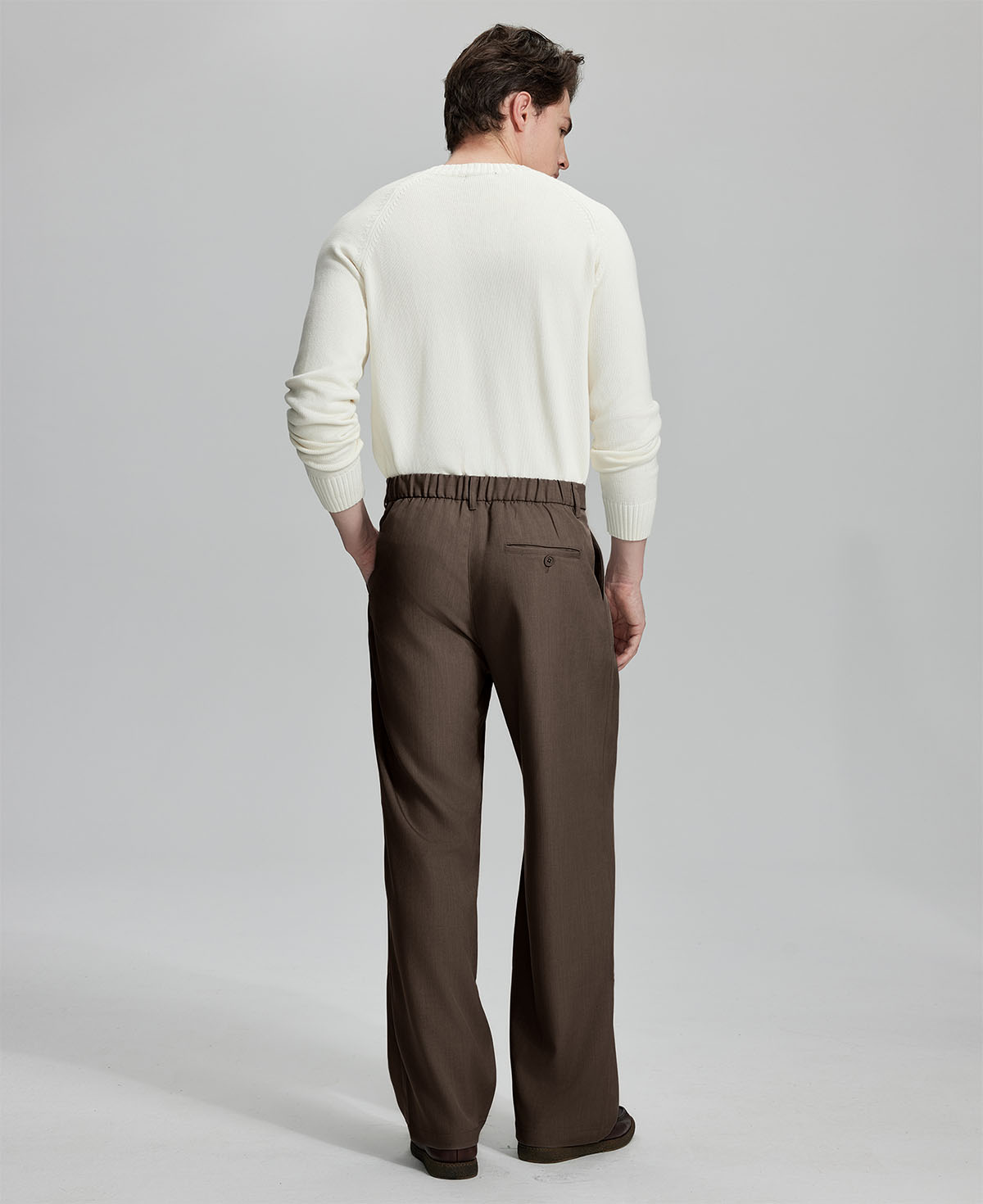 Side view of man adjusting cuff while highlighting the clean lines and casual elegance of coffee pleated baggy dress pants.