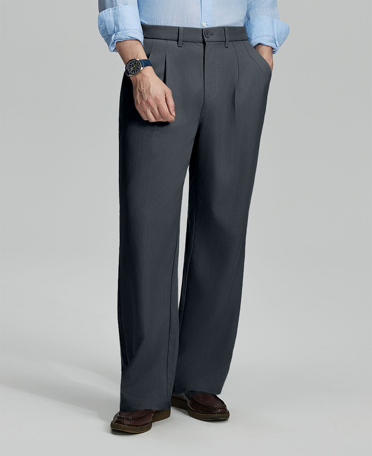 Man in light blue shirt, dark gray pleated pants, and brown shoes, hand in pocket showcasing relaxed fit and drape.