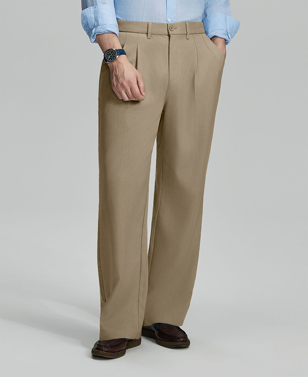 Man in light blue shirt, khaki pleated pants, and brown shoes, hand in pocket showcasing relaxed fit and drape.