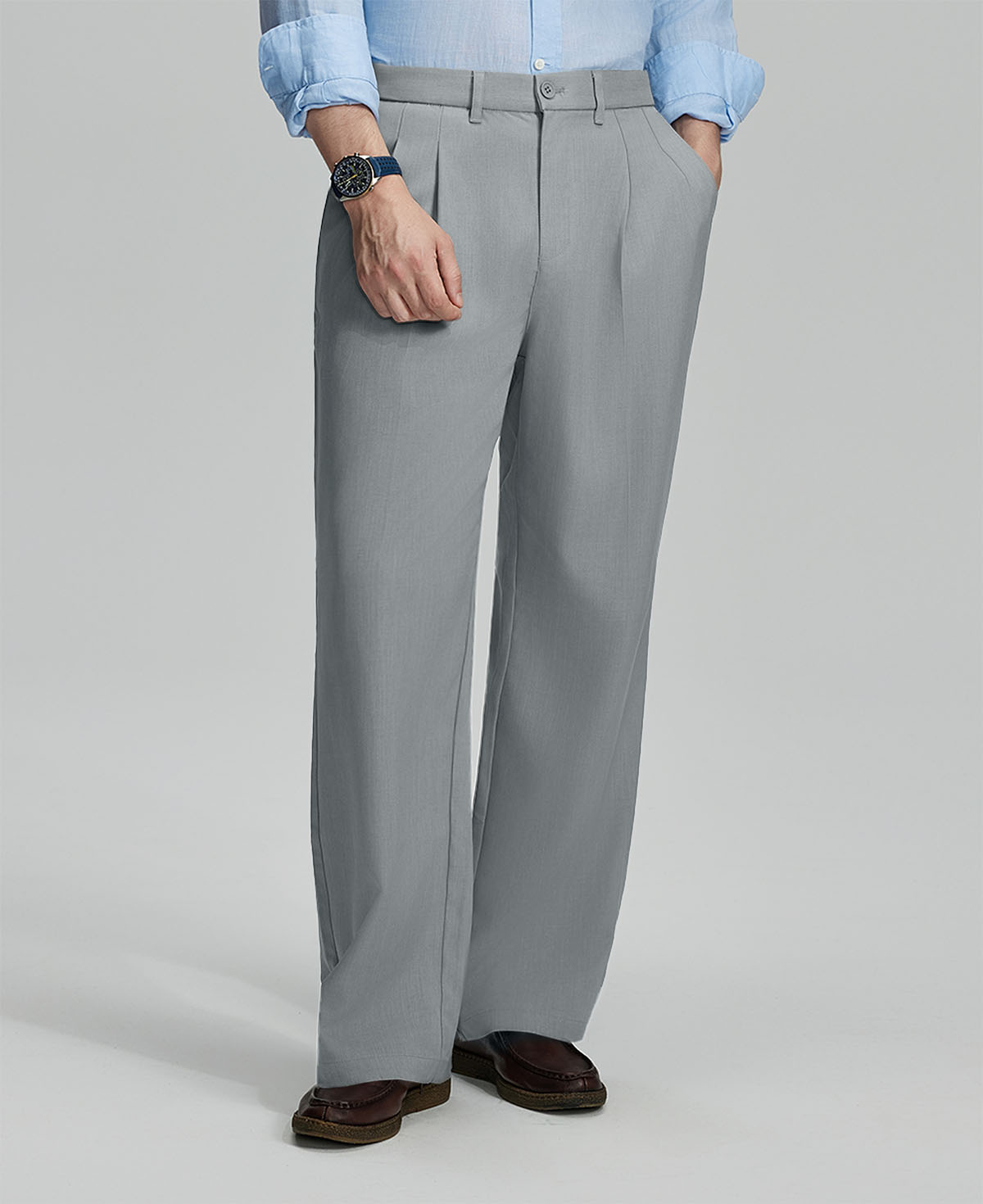 Man in light blue shirt, light grey pleated pants, and brown shoes, hand in pocket showcasing relaxed fit and drape.