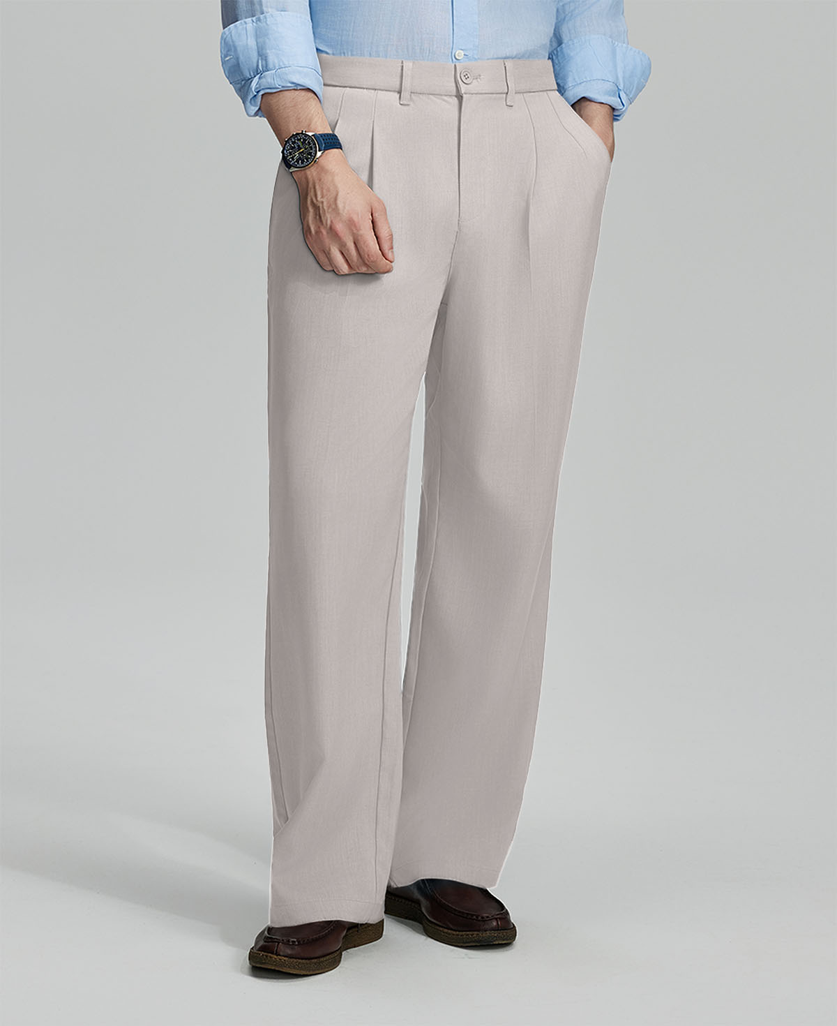 Man in light blue shirt, stone grey pleated pants, and brown shoes, hand in pocket showcasing relaxed fit and drape.
