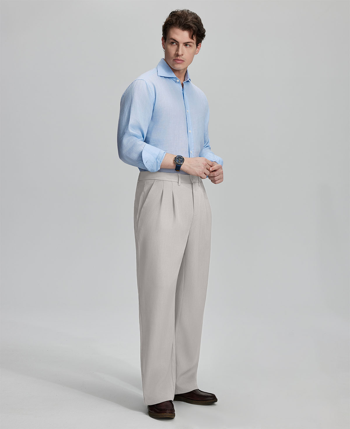 Side view of man adjusting cuff while highlighting the clean lines and casual elegance of stone grey pleated baggy dress pants.