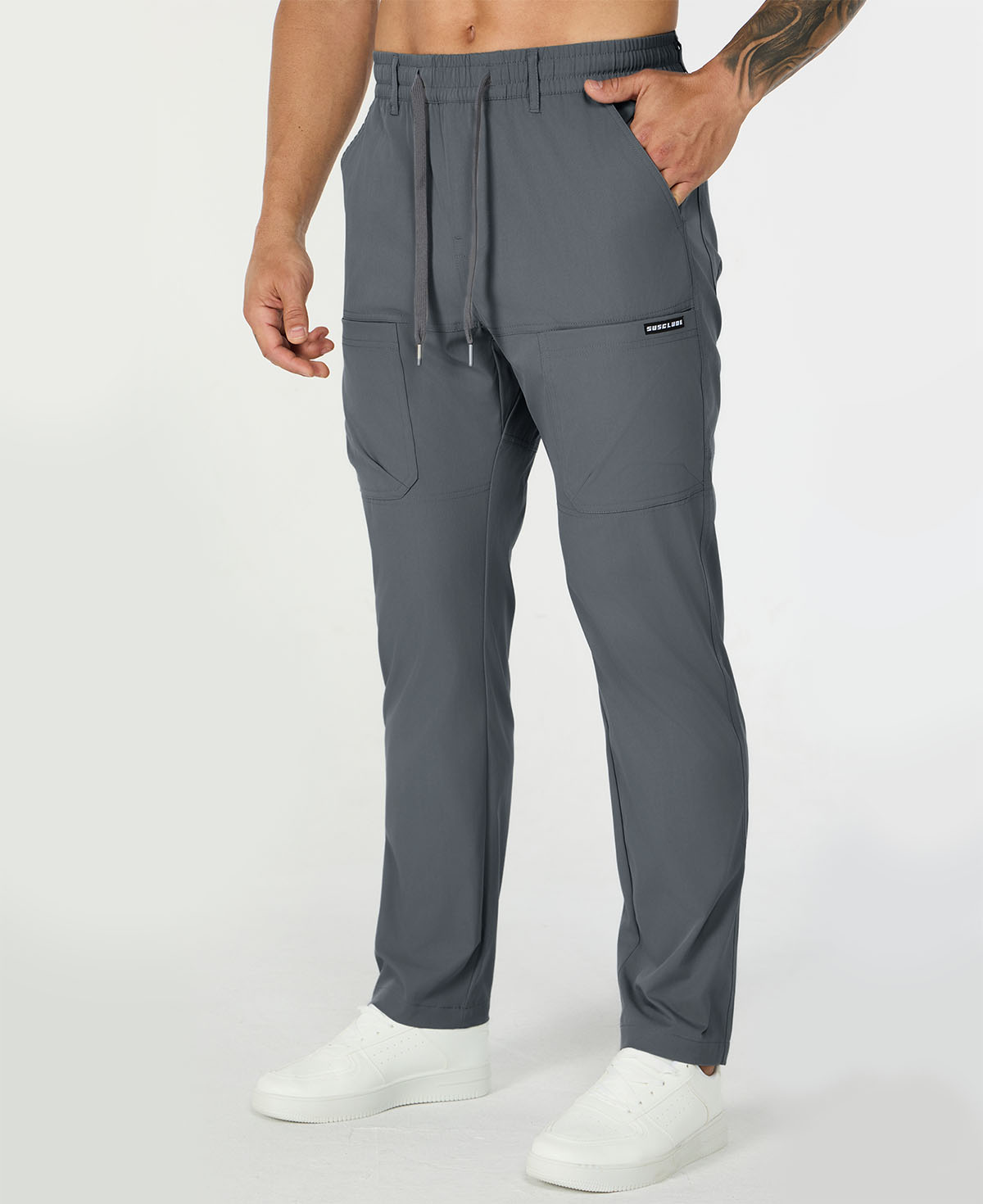 Man in dark grey multipants with drawstring waist and multiple pockets, white sneakers, hand in pocket showing side logo.