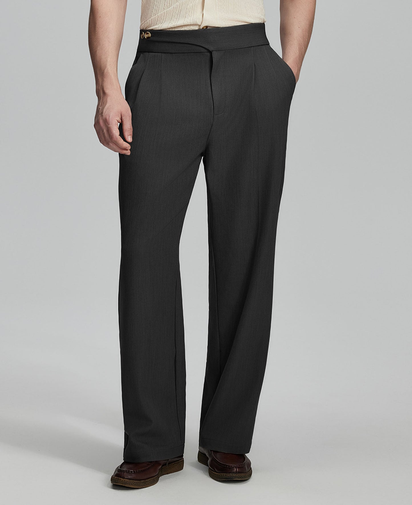 Man in beige sweater, black Gurkha pants & brown shoes, hand in pocket showing pleated wide leg drape and flow.