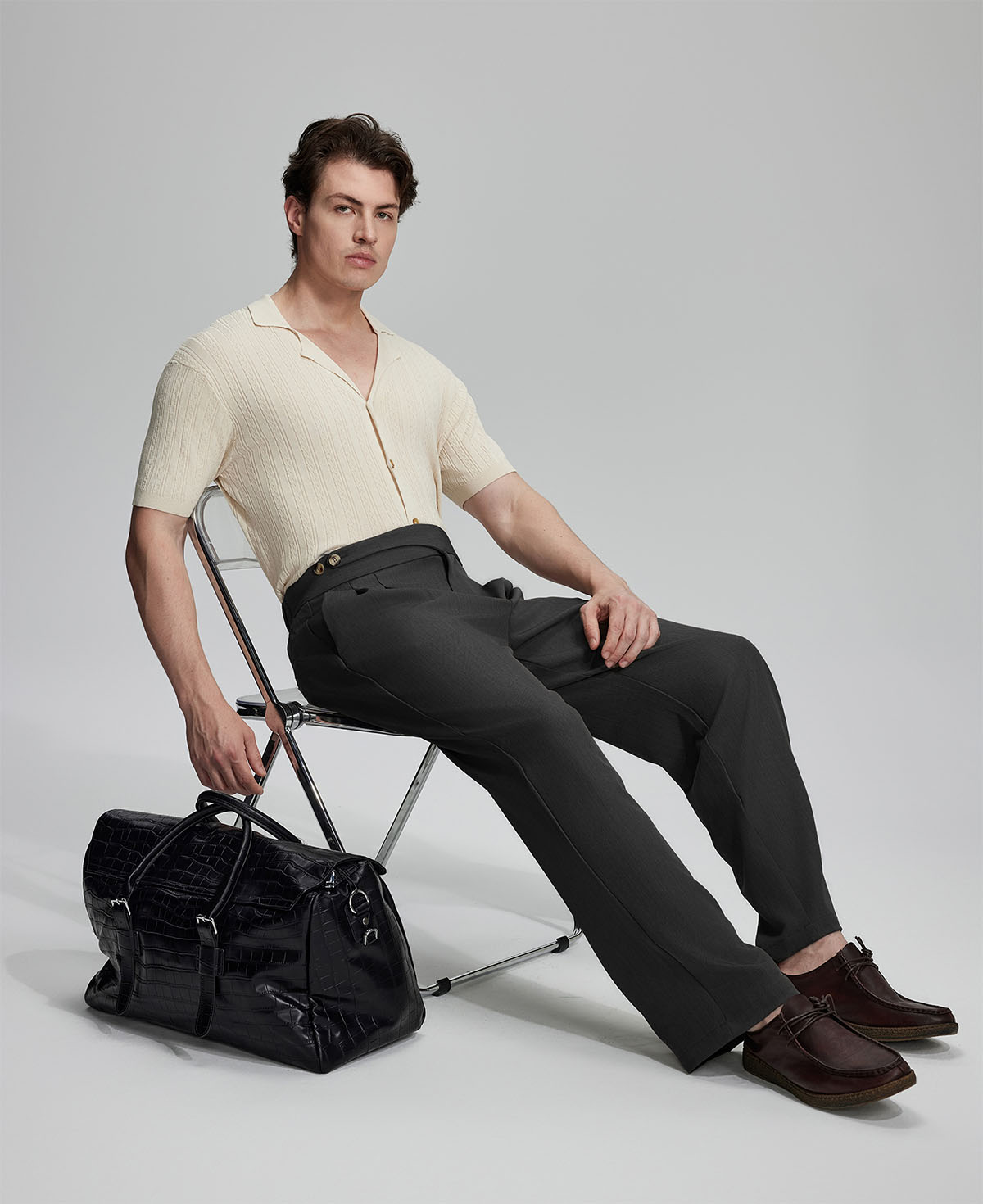 Model in beige knit and black Gurkha trousers with black bag, leaning on chair, elegant smart-casual look.