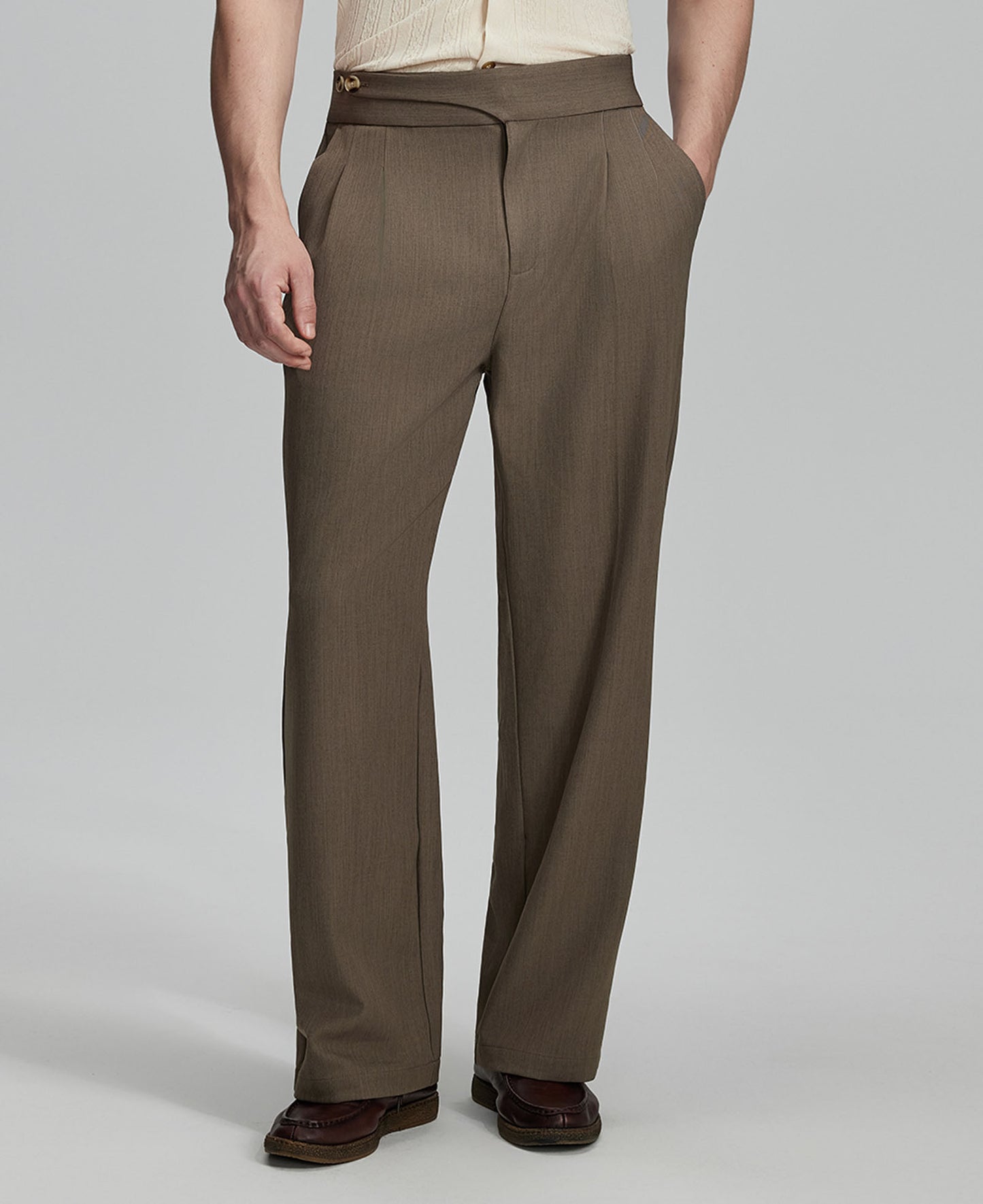 Man in beige sweater, brown Gurkha pants & brown shoes, hand in pocket showing pleated wide leg drape and flow.