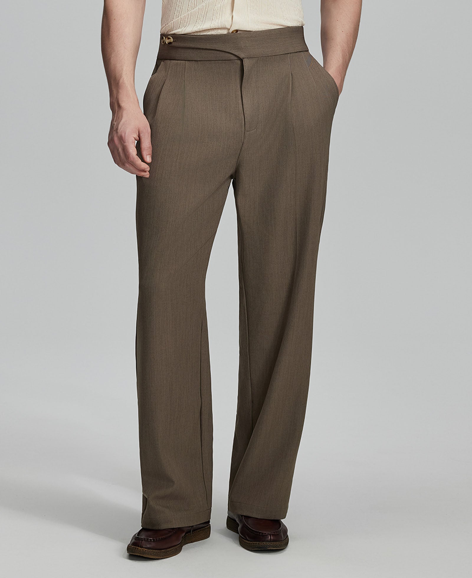 Man in beige sweater, brown Gurkha pants & brown shoes, hand in pocket showing pleated wide leg drape and flow.