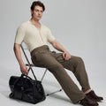 Model in beige knit and brown Gurkha trousers with black bag, leaning on chair, elegant smart-casual look.