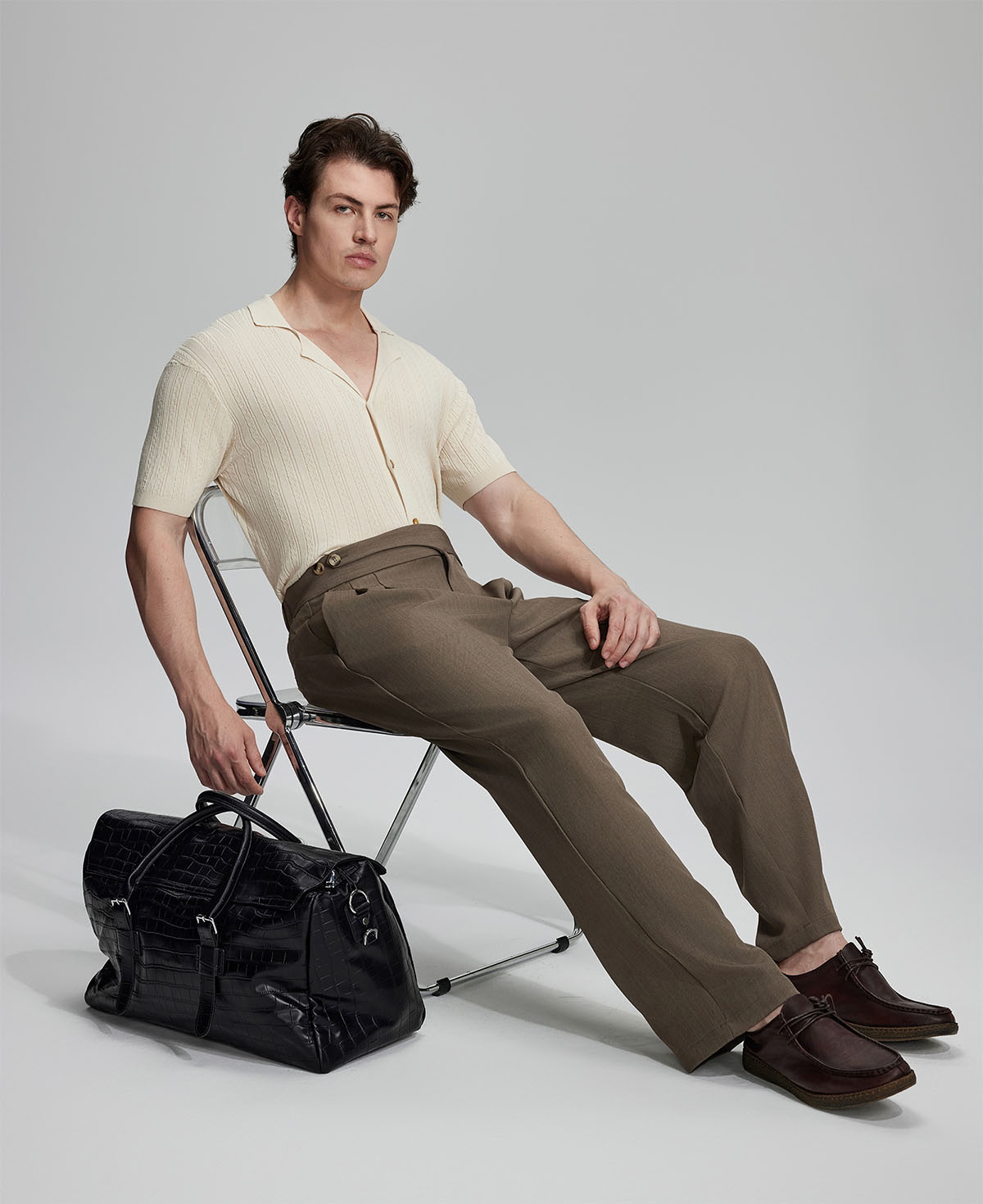 Model in beige knit and brown Gurkha trousers with black bag, leaning on chair, elegant smart-casual look.