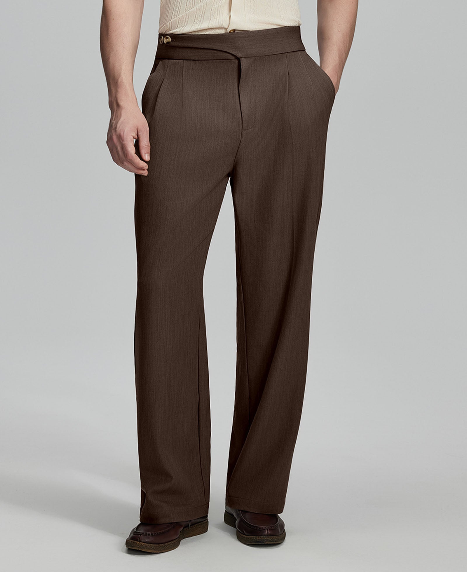Man in beige sweater, brown Gurkha pants & brown shoes, hand in pocket showing pleated wide leg drape and flow.