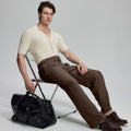Model in beige knit and brown Gurkha trousers with black bag, leaning on chair, elegant smart-casual look.