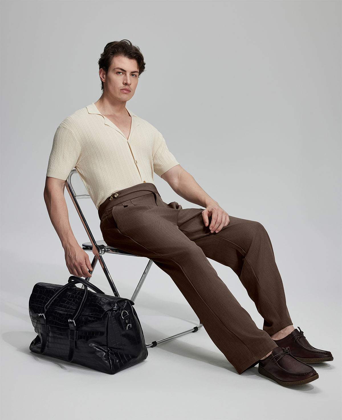 Model in beige knit and brown Gurkha trousers with black bag, leaning on chair, elegant smart-casual look.