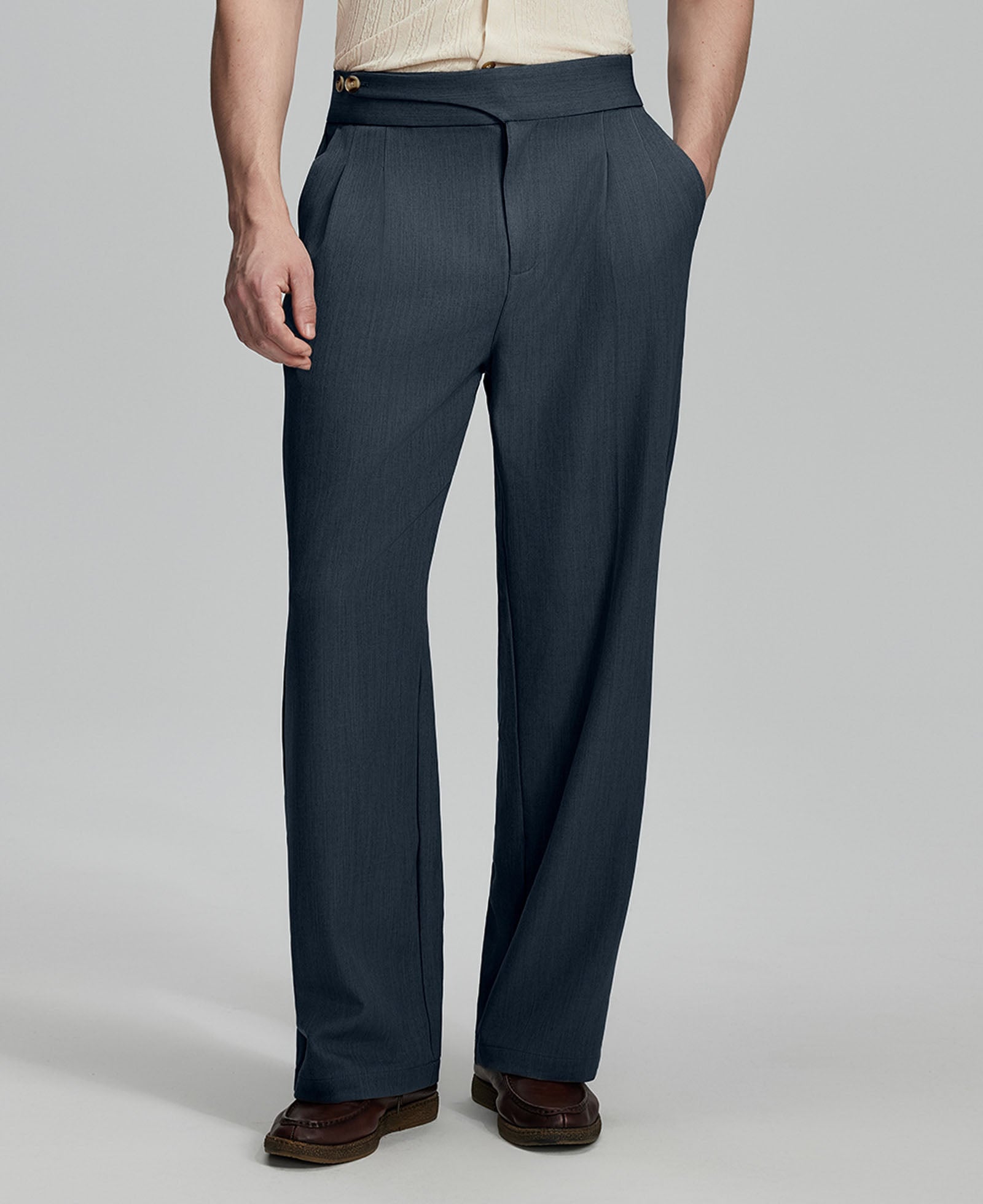 Man in beige sweater, grey Gurkha pants & brown shoes, hand in pocket showing pleated wide leg drape and flow.