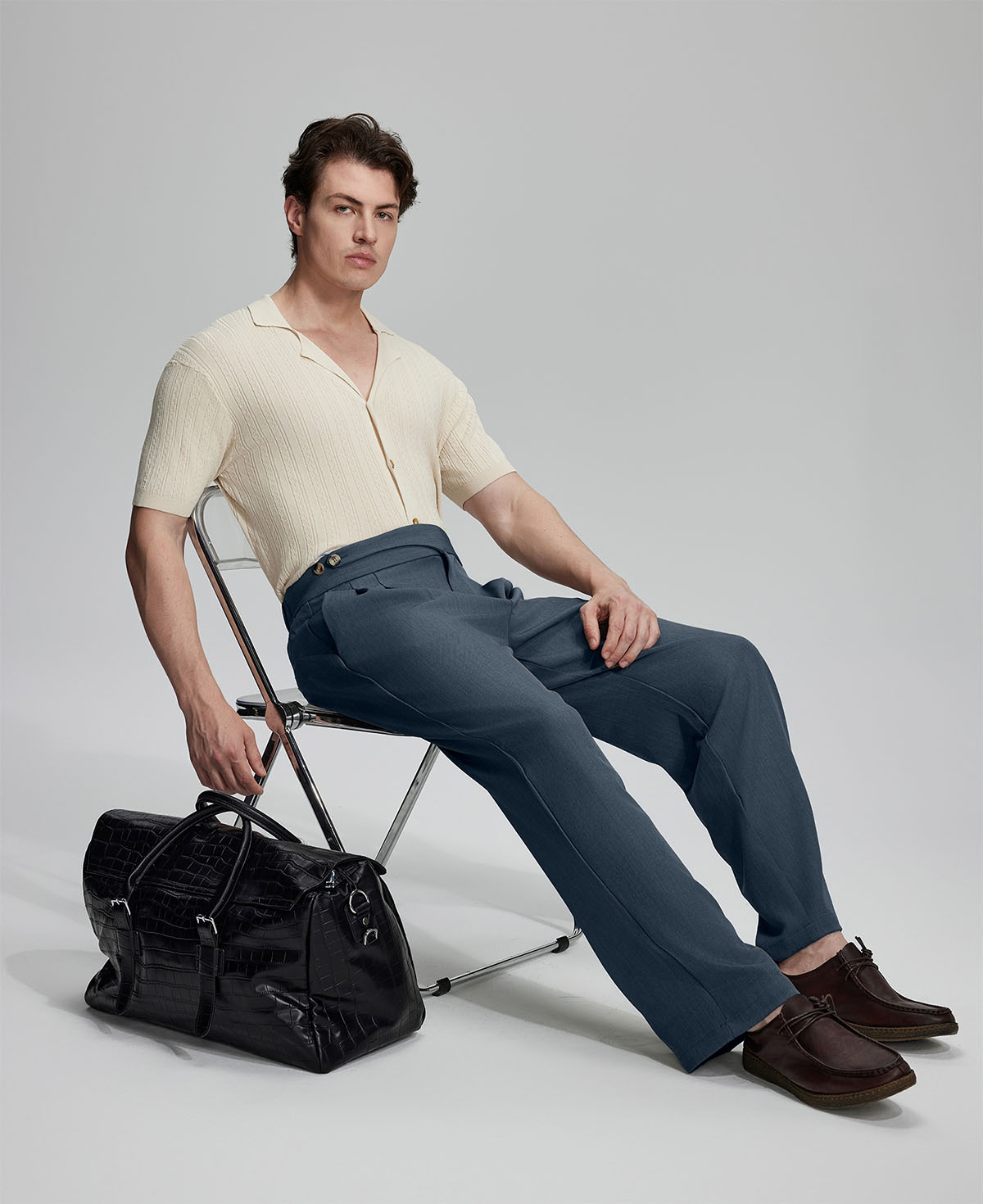 Model in beige knit and grey Gurkha trousers with black bag, leaning on chair, elegant smart-casual look.