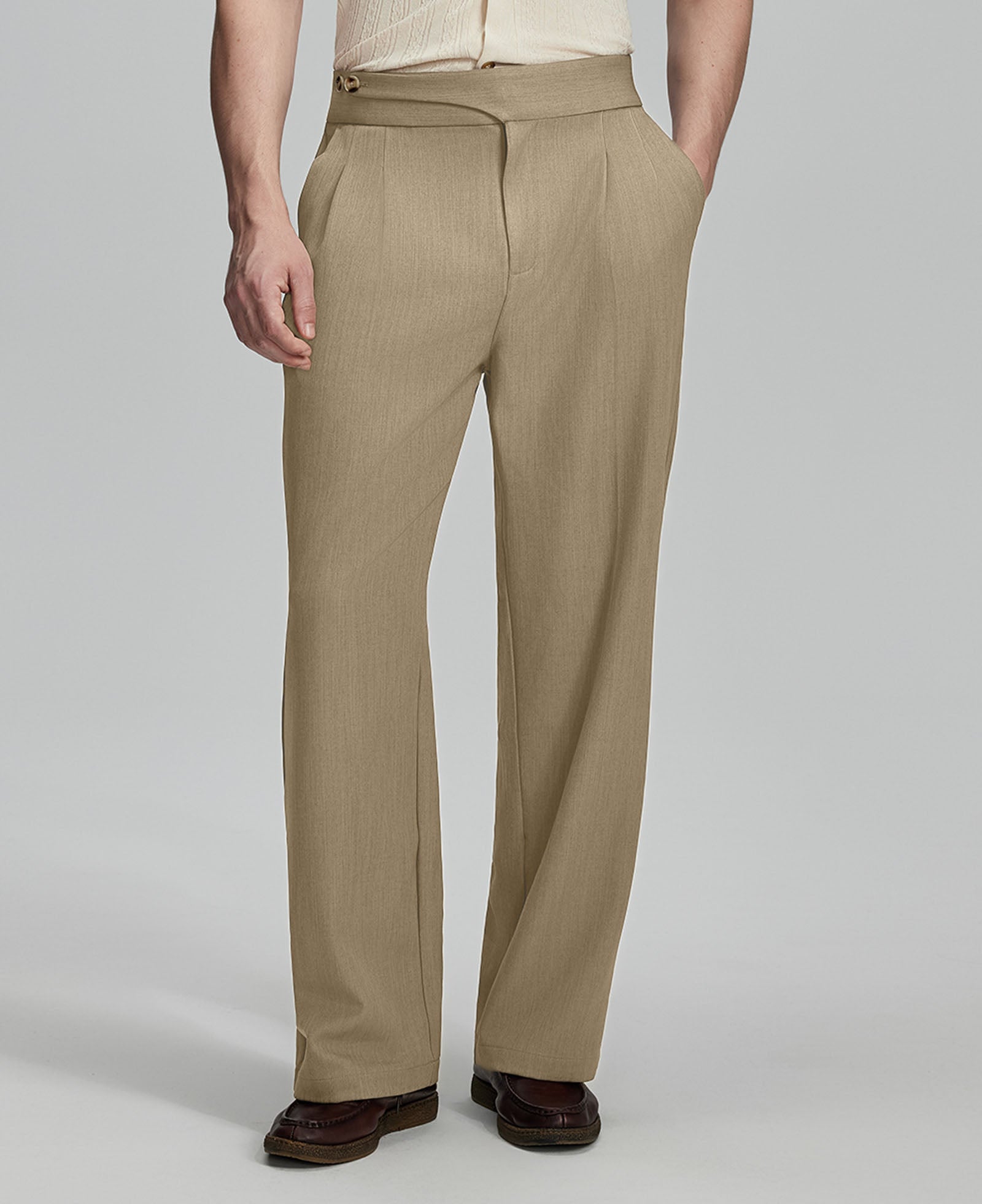 Man in beige sweater, beige Gurkha pants & brown shoes, hand in pocket showing pleated wide leg drape and flow.