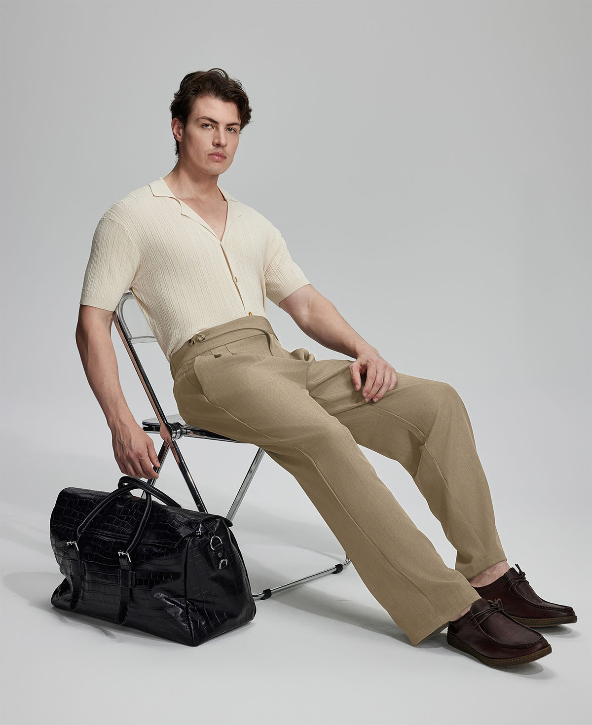 Model in beige knit and beige Gurkha trousers with black bag, leaning on chair, elegant smart-casual look.