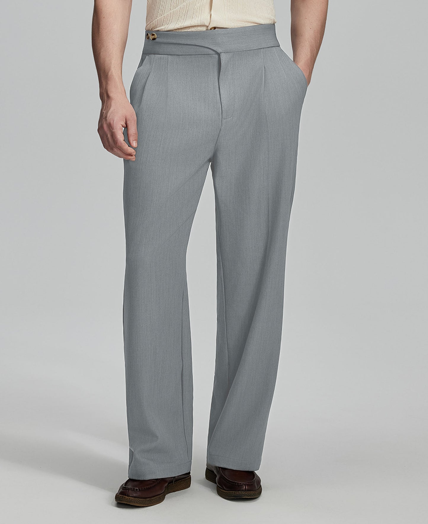 Man in beige sweater, grey Gurkha pants & brown shoes, hand in pocket showing pleated wide leg drape and flow.