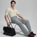 Model in beige knit and grey Gurkha trousers with black bag, leaning on chair, elegant smart-casual look.