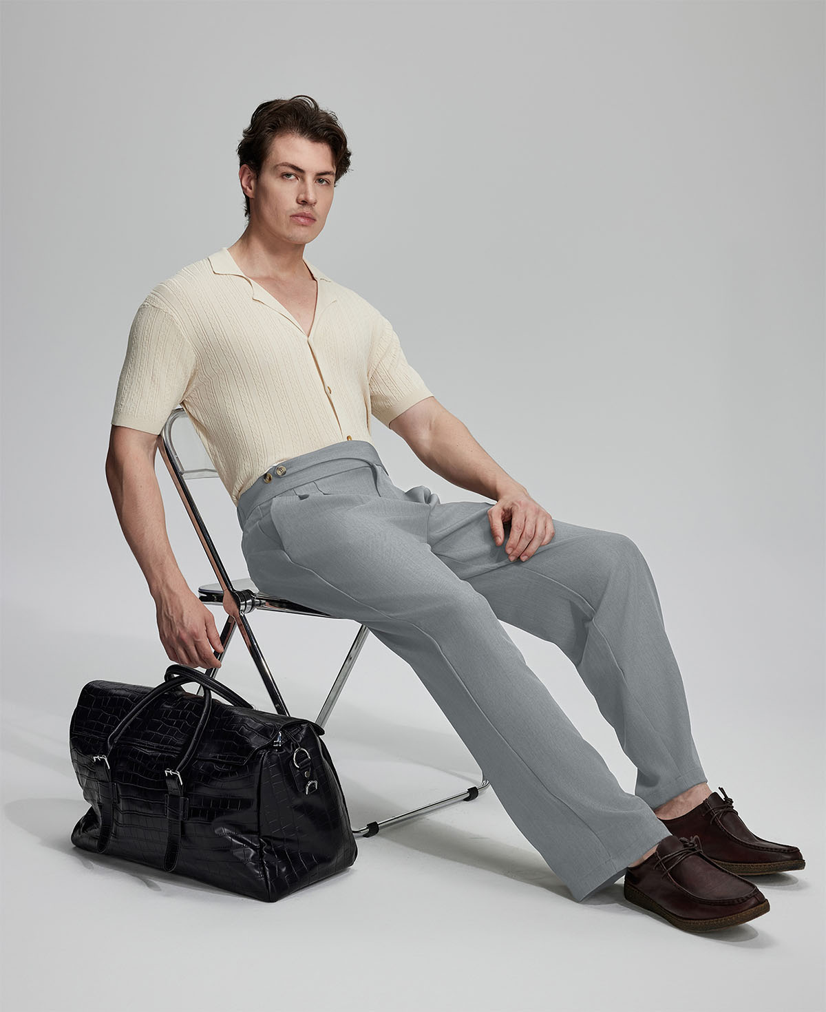 Model in beige knit and grey Gurkha trousers with black bag, leaning on chair, elegant smart-casual look.