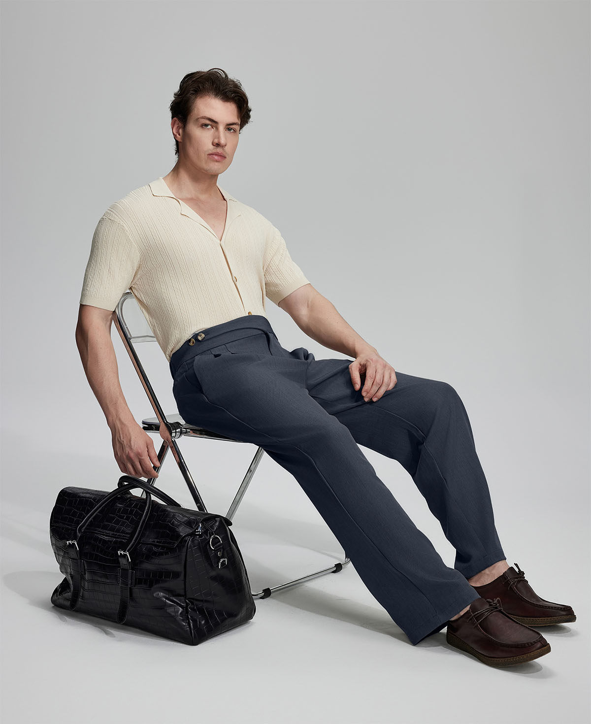 Model in beige knit and navy Gurkha trousers with black bag, leaning on chair, elegant smart-casual look.