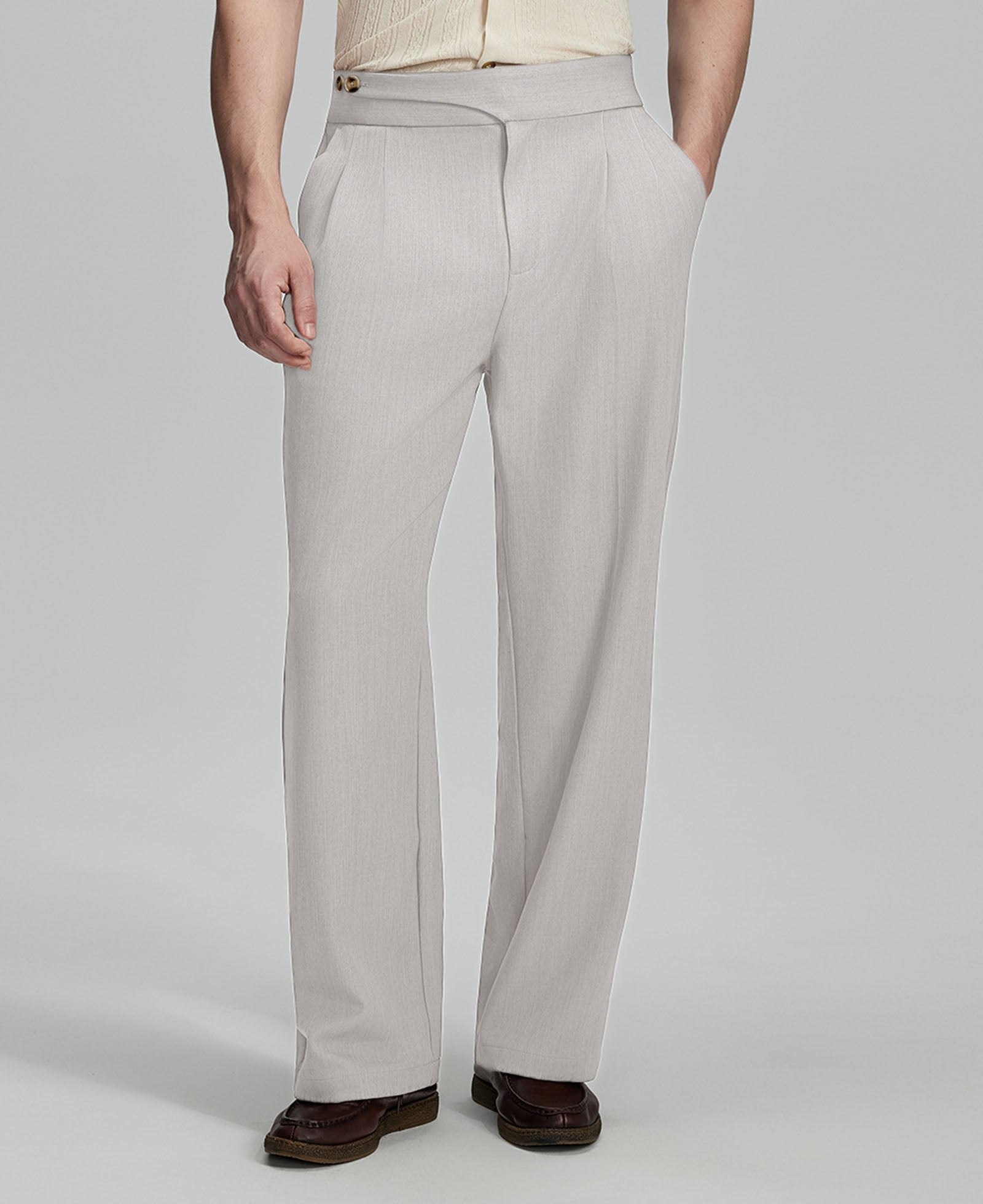Man in beige sweater, grey Gurkha pants & brown shoes, hand in pocket showing pleated wide leg drape and flow.
