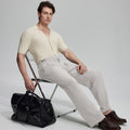 Model in beige knit and grey Gurkha trousers with black bag, leaning on chair, elegant smart-casual look.
