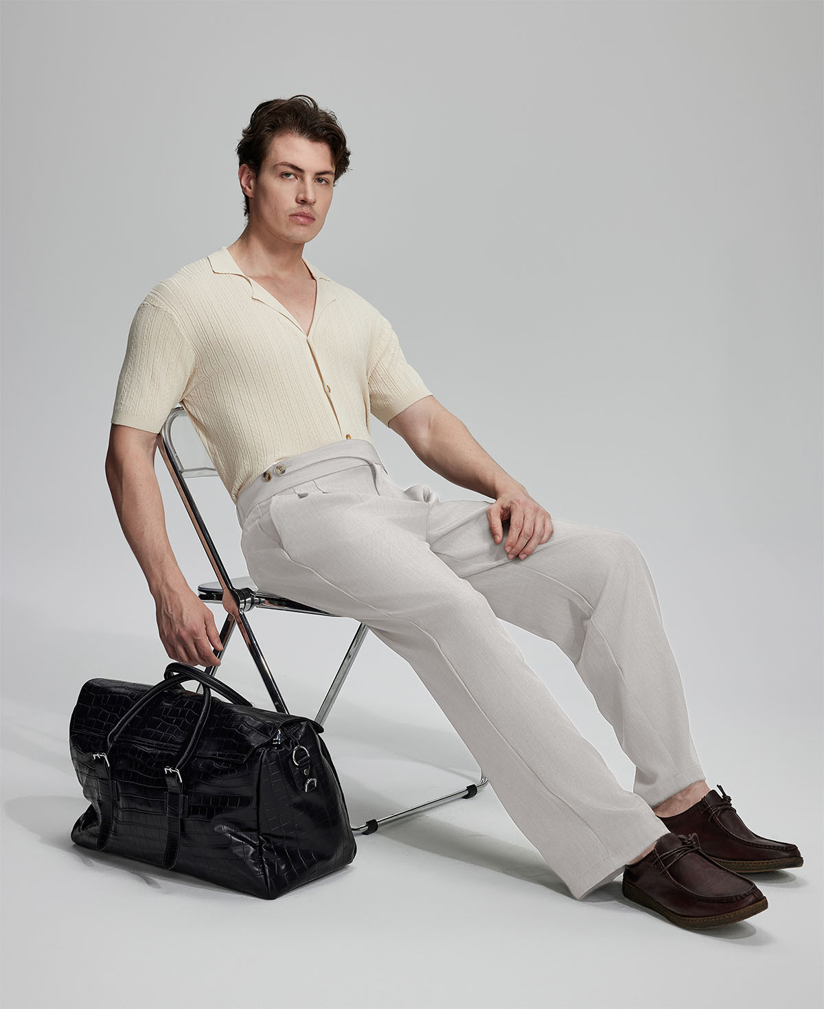 Model in beige knit and grey Gurkha trousers with black bag, leaning on chair, elegant smart-casual look.