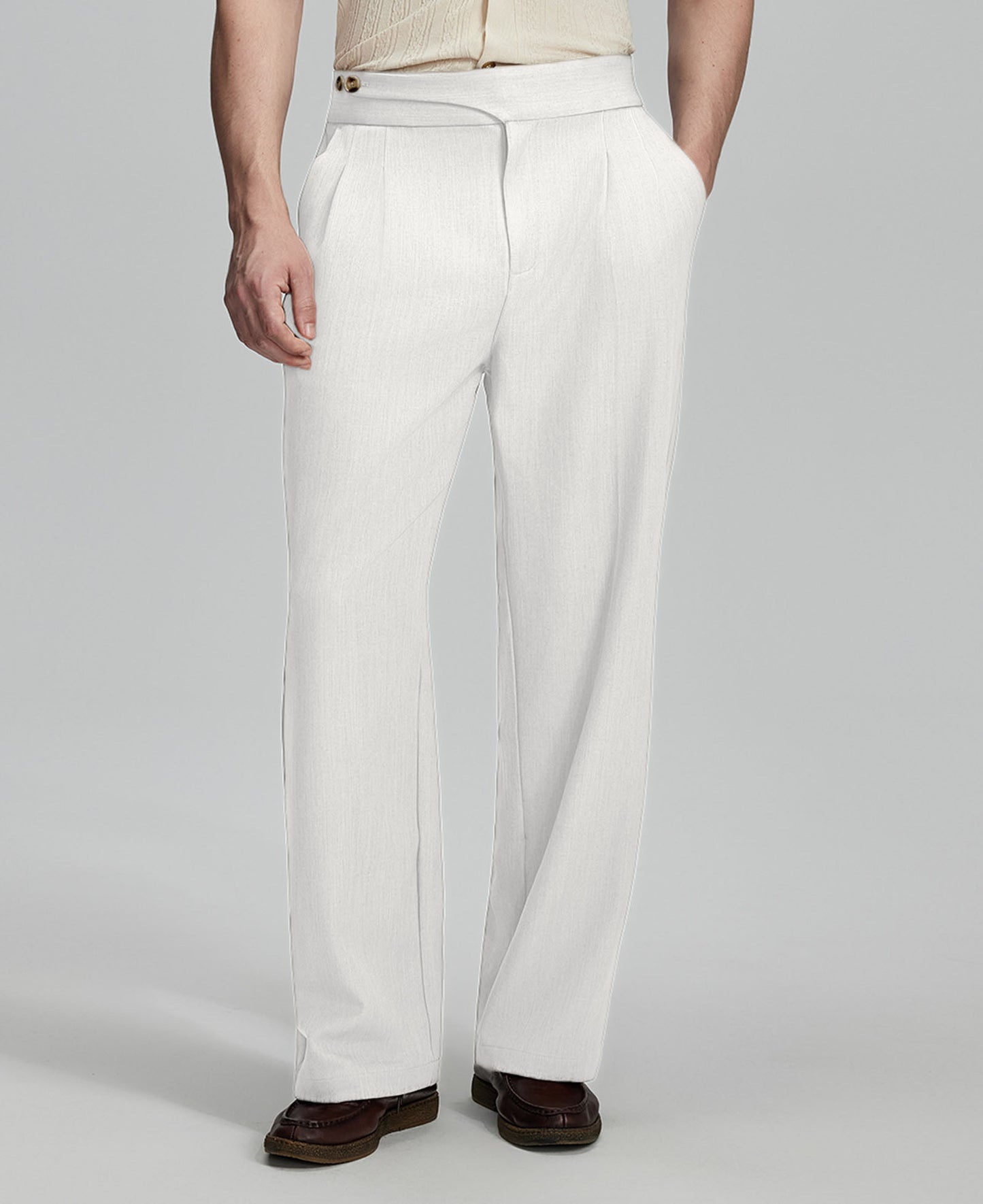Man in beige sweater, white Gurkha pants & brown shoes, hand in pocket showing pleated wide leg drape and flow.
