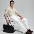 Model in beige knit and white Gurkha trousers with black bag, leaning on chair, elegant smart-casual look.