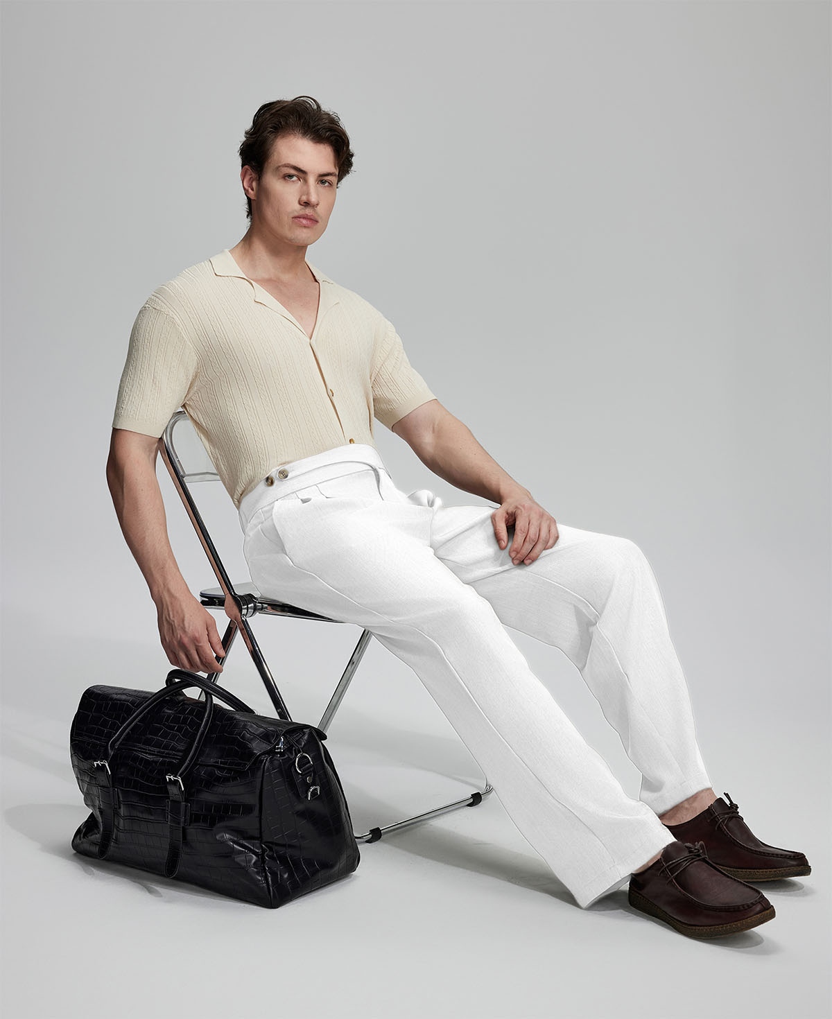 Model in beige knit and white Gurkha trousers with black bag, leaning on chair, elegant smart-casual look.