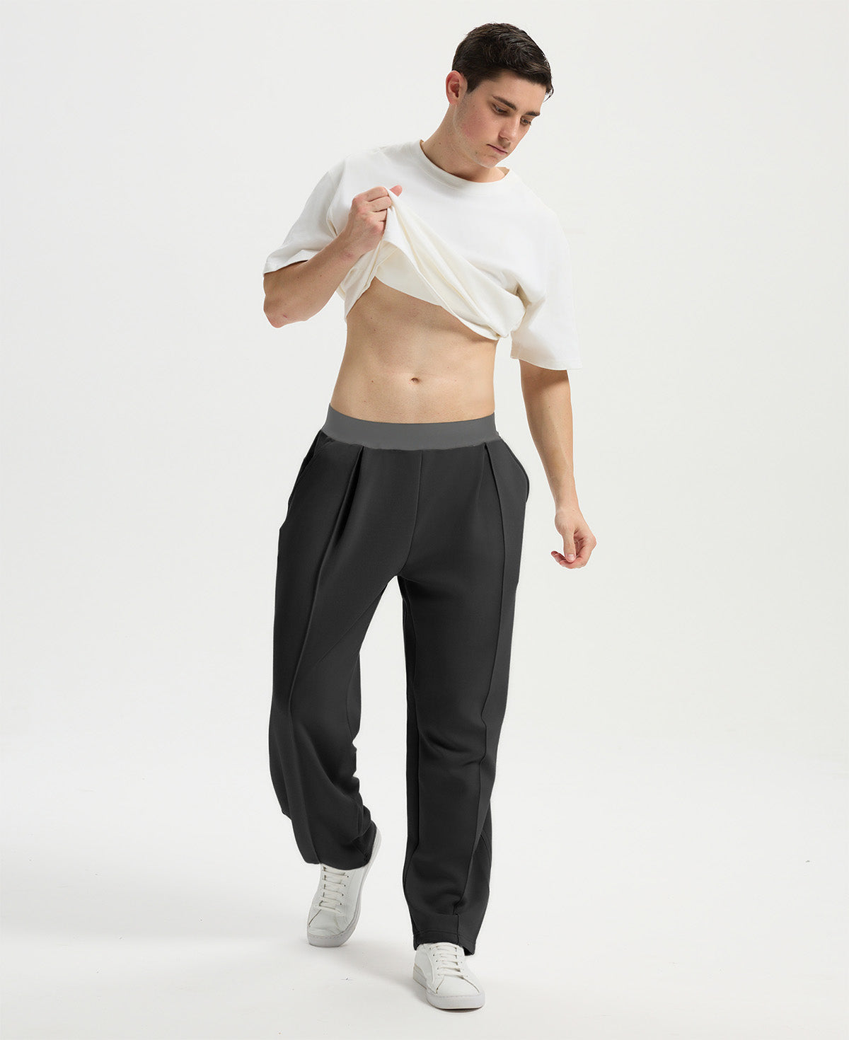 Man wearing white tee and black men's pleated baggy sweatpants, lifting shirt to show waist detail and fit.