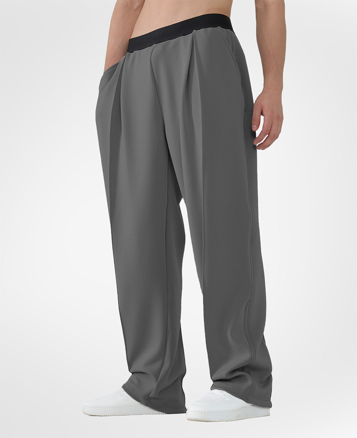 Man in dark grey pleated baggy sweatpants with white shoes, hand in pocket, showcasing wide leg and contrasting elastic waist.
