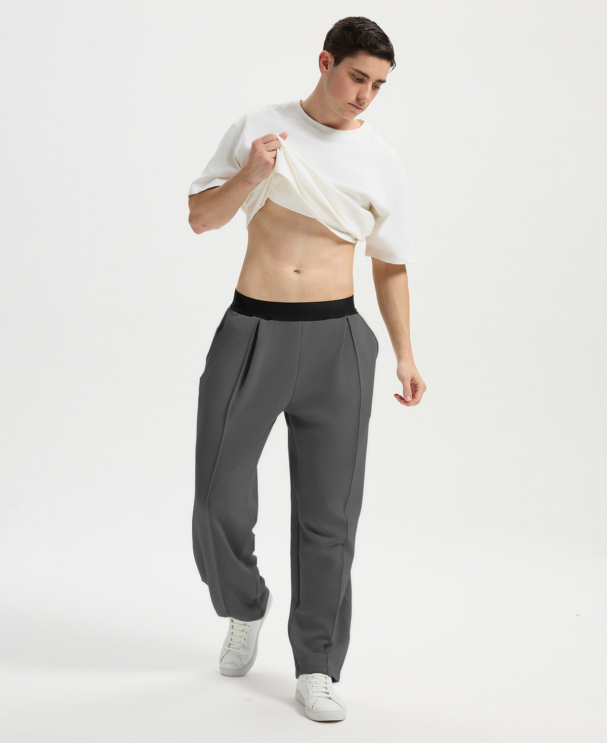 Man wearing white tee and dark grey men's pleated baggy sweatpants, lifting shirt to show waist detail and fit.