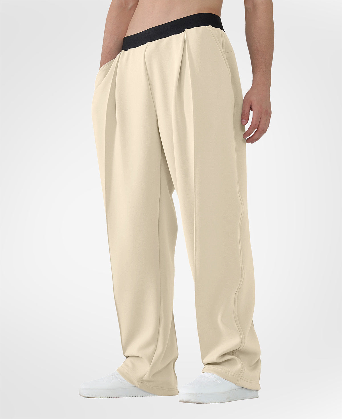 Man in khaki pleated baggy sweatpants with white shoes, hand in pocket, showcasing wide leg and contrasting elastic waist.