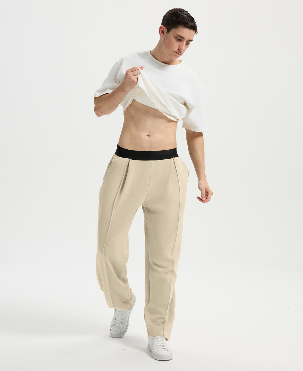 Man wearing white tee and khaki men's pleated baggy sweatpants, lifting shirt to show waist detail and fit.