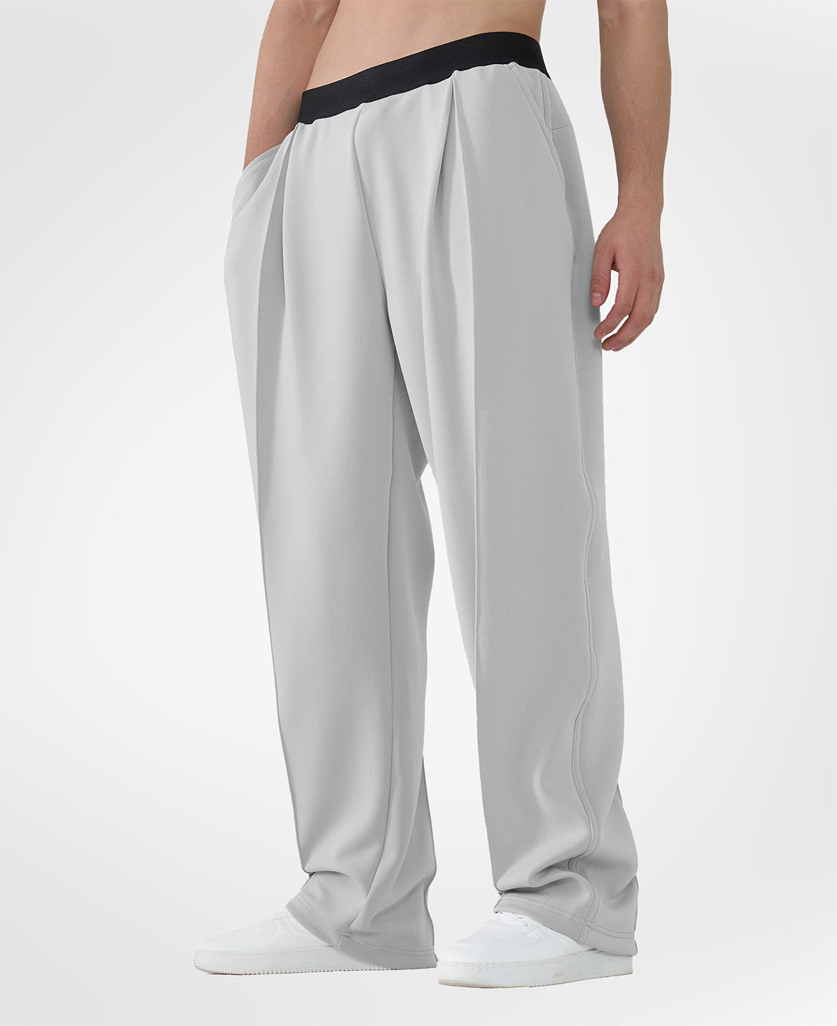Man in light grey pleated baggy sweatpants with white shoes, hand in pocket, showcasing wide leg and contrasting elastic waist.