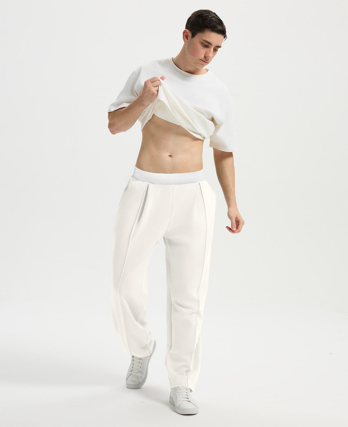Man wearing white tee and white men's pleated baggy sweatpants, lifting shirt to show waist detail and fit.