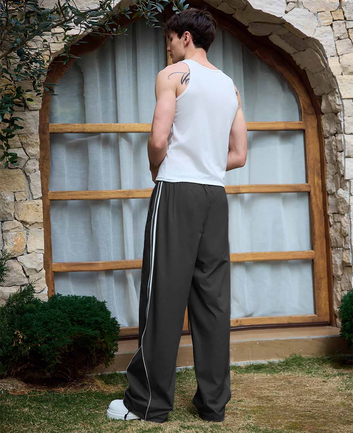 Man in black baggy wide leg sweatpants, white thick-soled shoes, back to camera, side stance on grass, showing loose fit and stripes.