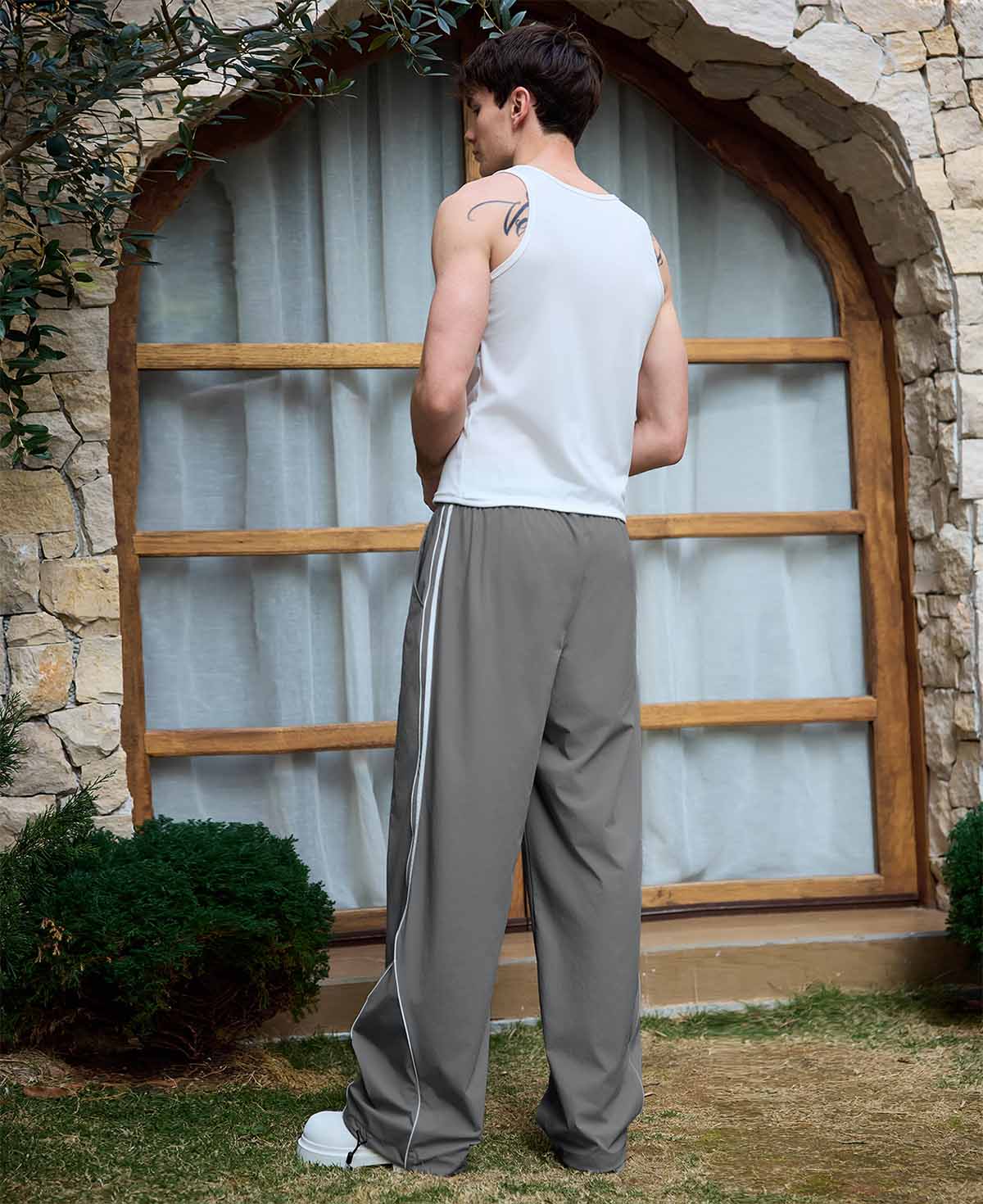 Man in dark grey baggy wide leg sweatpants, white thick-soled shoes, back to camera, side stance on grass, showing loose fit and stripes.
