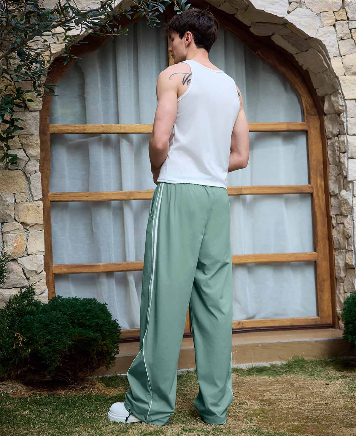 Man in green baggy wide leg sweatpants, white thick-soled shoes, back to camera, side stance on grass, showing loose fit and stripes.