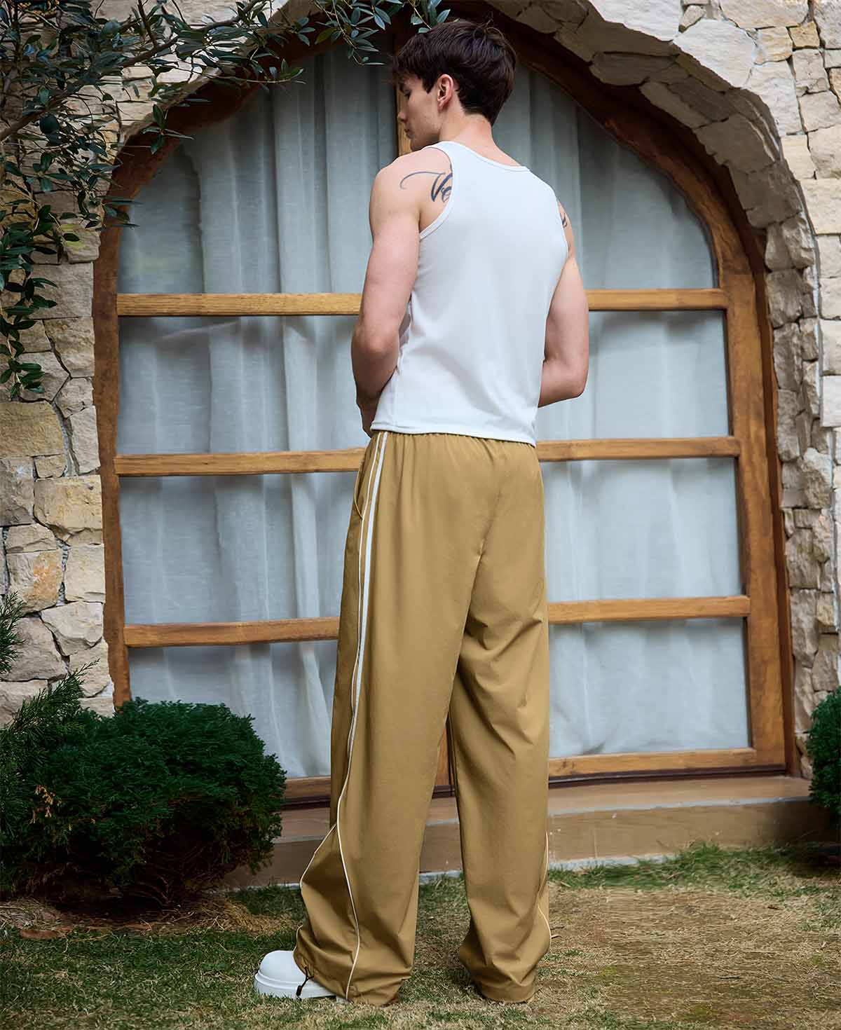 Man in khaki baggy wide leg sweatpants, white thick-soled shoes, back to camera, side stance on grass, showing loose fit and stripes.